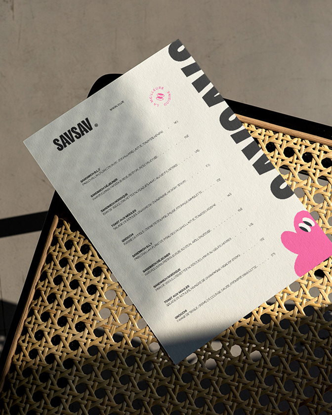 Un menu papier SavSav est posé sur une table extérieure en métal avec motif graphique rose.