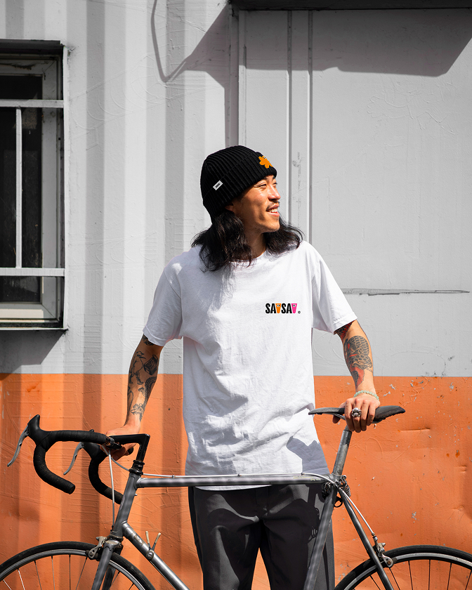 Un portrait lifestyle SavSav montre un homme en t-shirt blanc et bonnet noir tenant un vélo devant un mur orange et blanc.