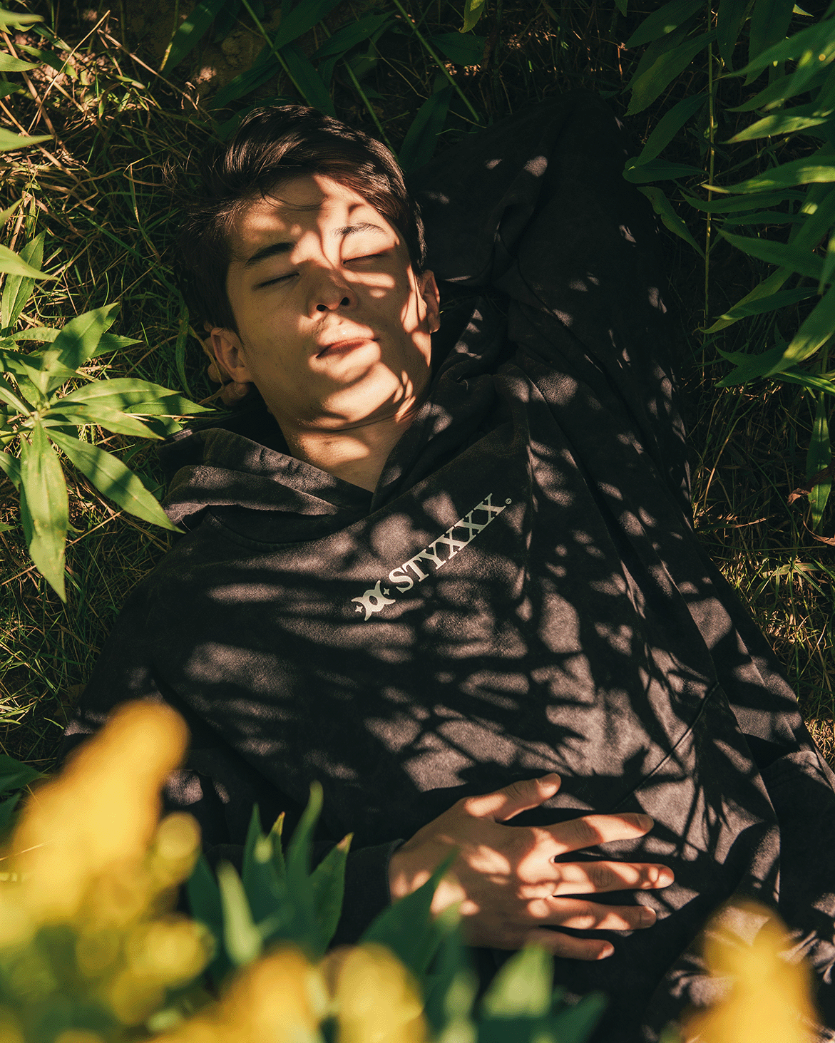 Young man lying on grass surrounded by green plants and yellow flowers, with shadows casting over his face and black hoodie.