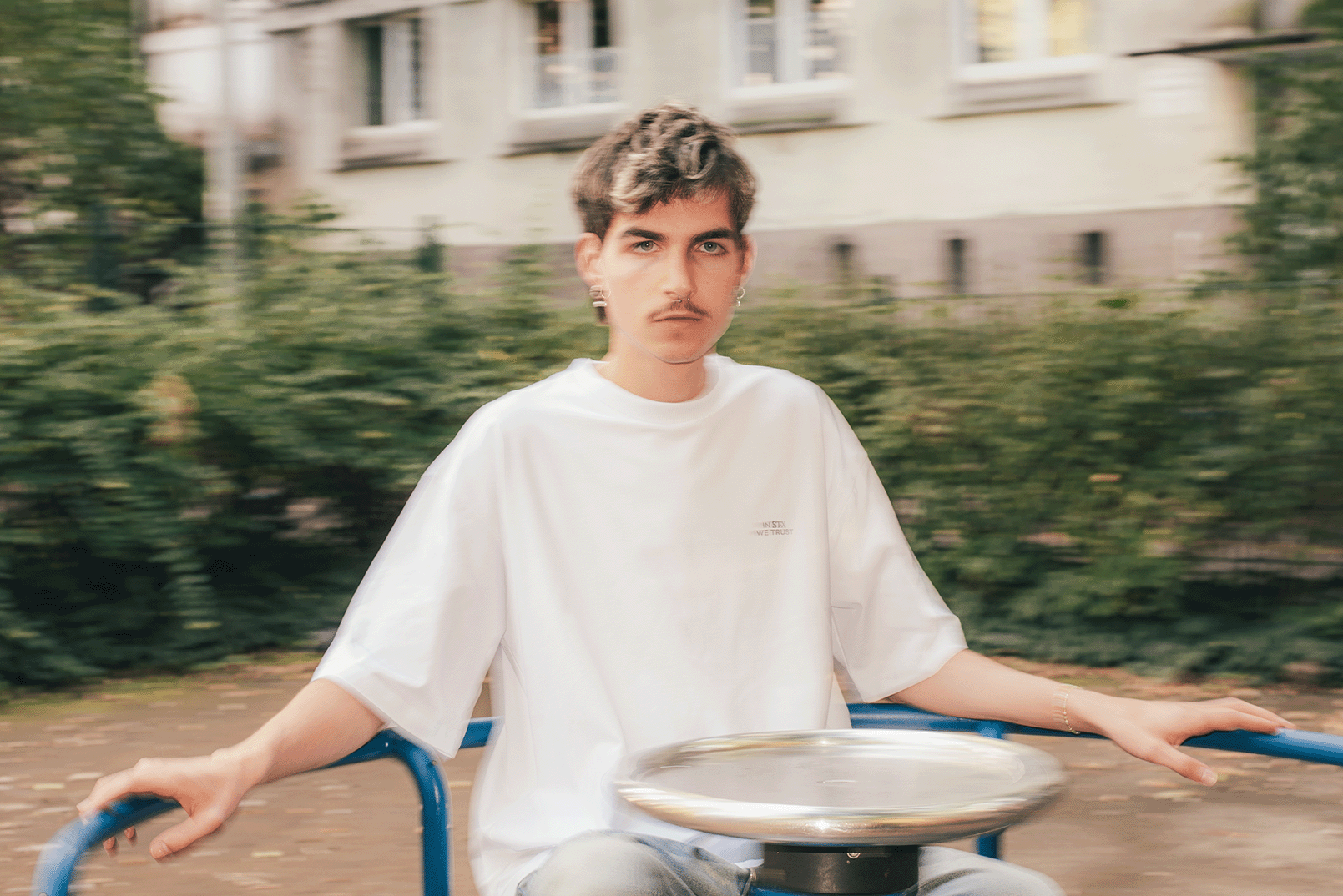 Young person with short hair and earrings sitting on blue playground equipment, holding onto bars with a blurred background of green bushes and building.