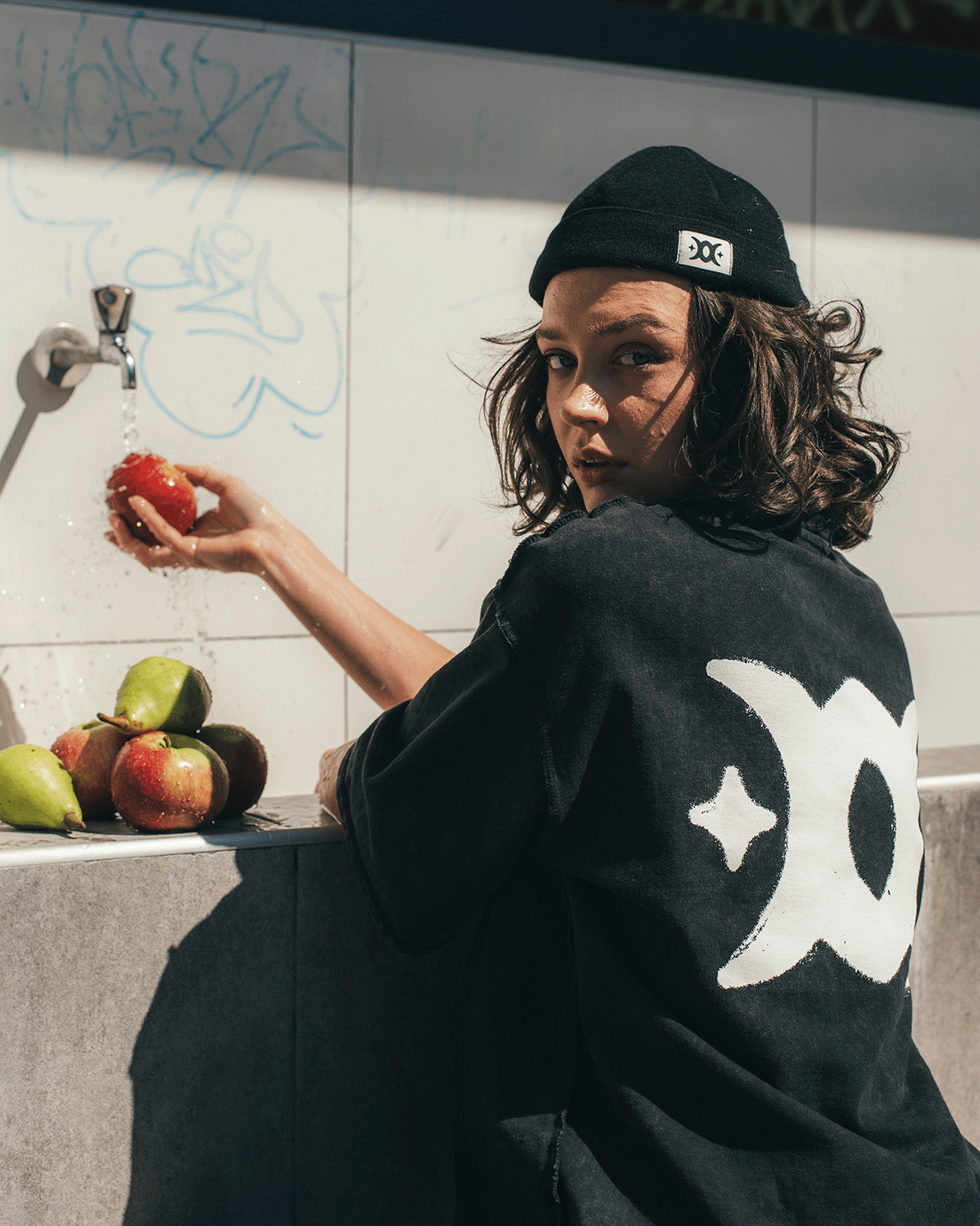 Person in a black beanie and black shirt washing a red apple under running water from a wall tap with pears and apples nearby.