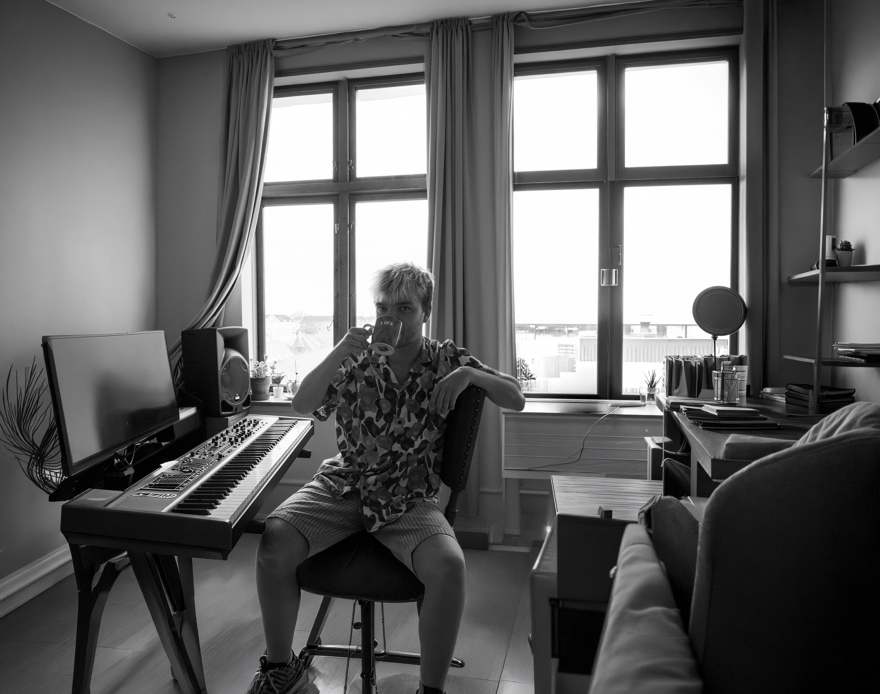 Ung mand sidder på en stol i et værelse med et keyboard foran sig og drikker kaffe.