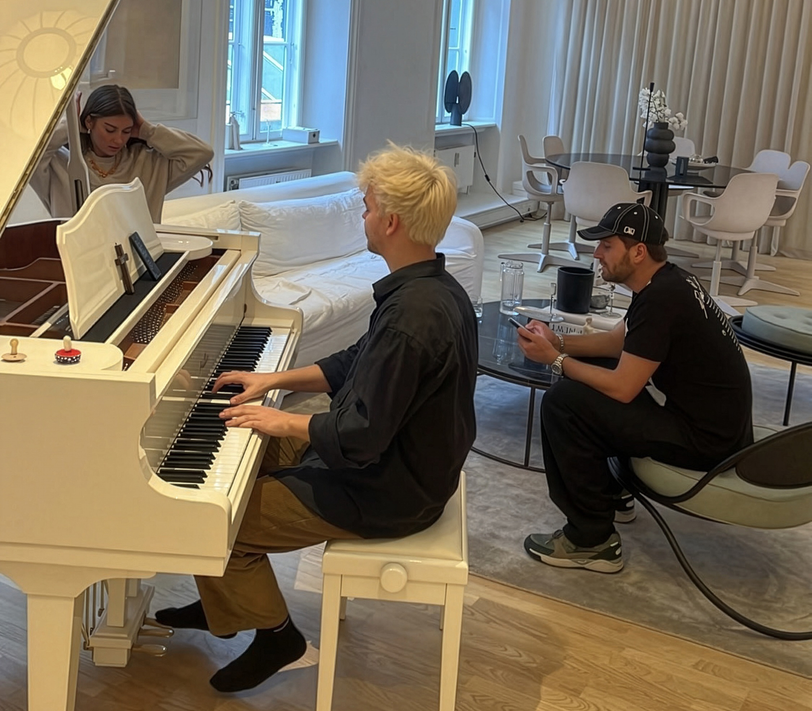 En mand med blondt hår spiller på et hvidt flygel, mens en kvinde sidder ved siden af og en anden mand i sort tøj ser på fra en stol med mobiltelefon.