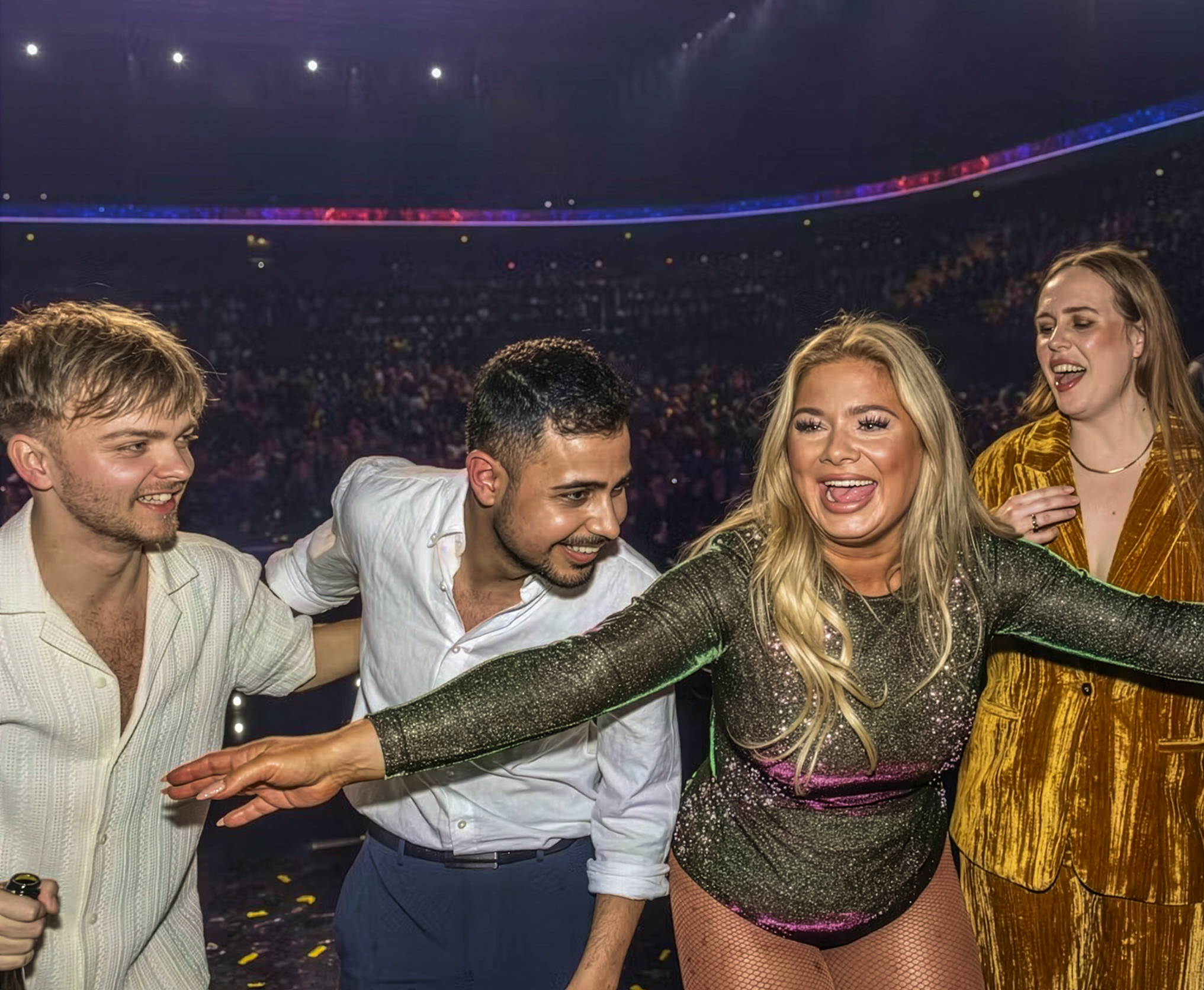 Fire glade personer, herunder en kvinde i glitrende grønt og pink tøj med mikrofon, poserer energisk foran et publikum og et kamerateam.