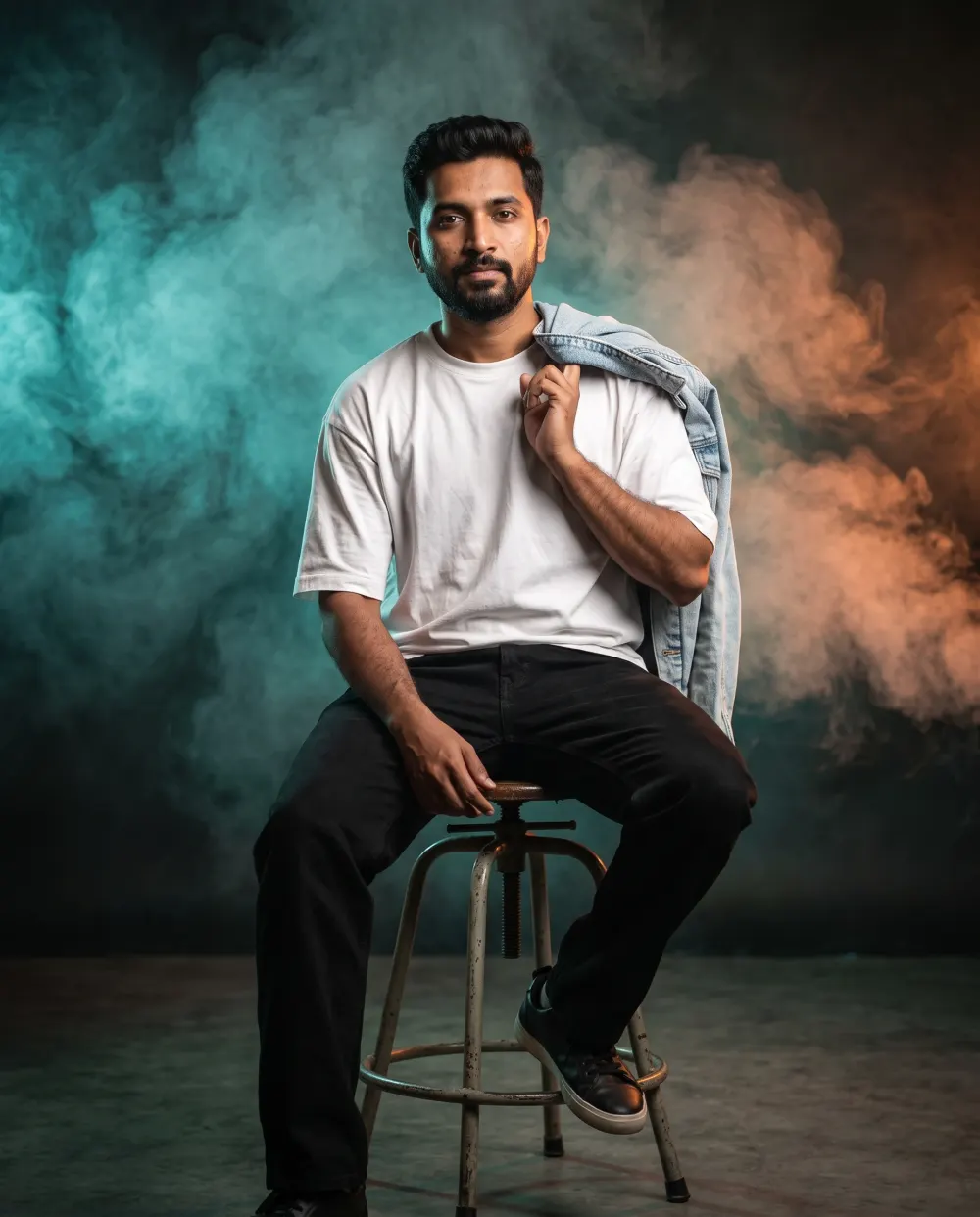 Man in a white tee on a metal stool with teal and orange smoke behind him