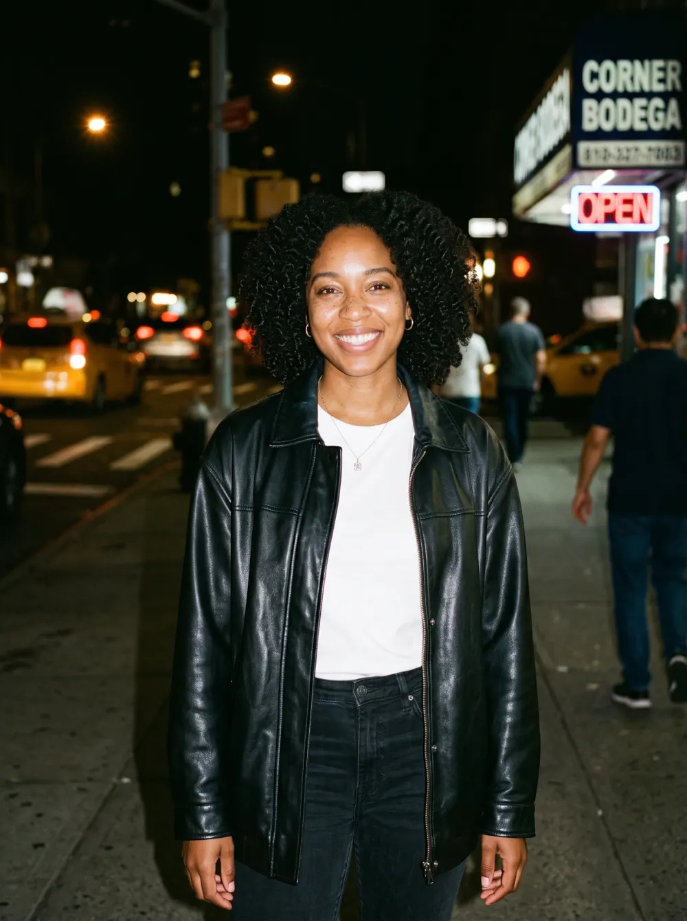 Flash-lit woman in a black leather jacket on a night sidewalk, corner bodega sign behind
