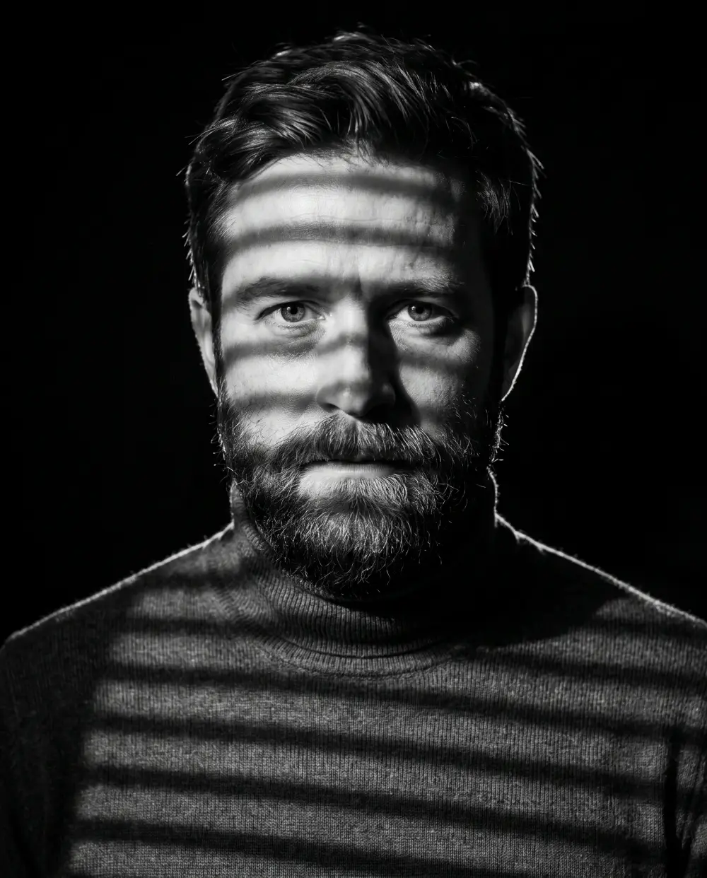 Black-and-white noir portrait of a bearded man with window-blind shadows across his face