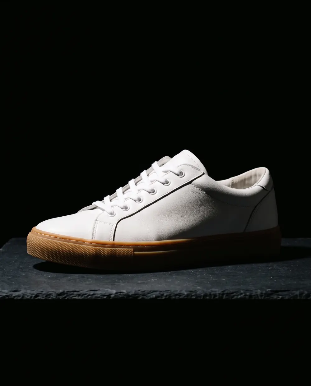 White leather sneaker with a gum sole on a dark slate block under one spotlight