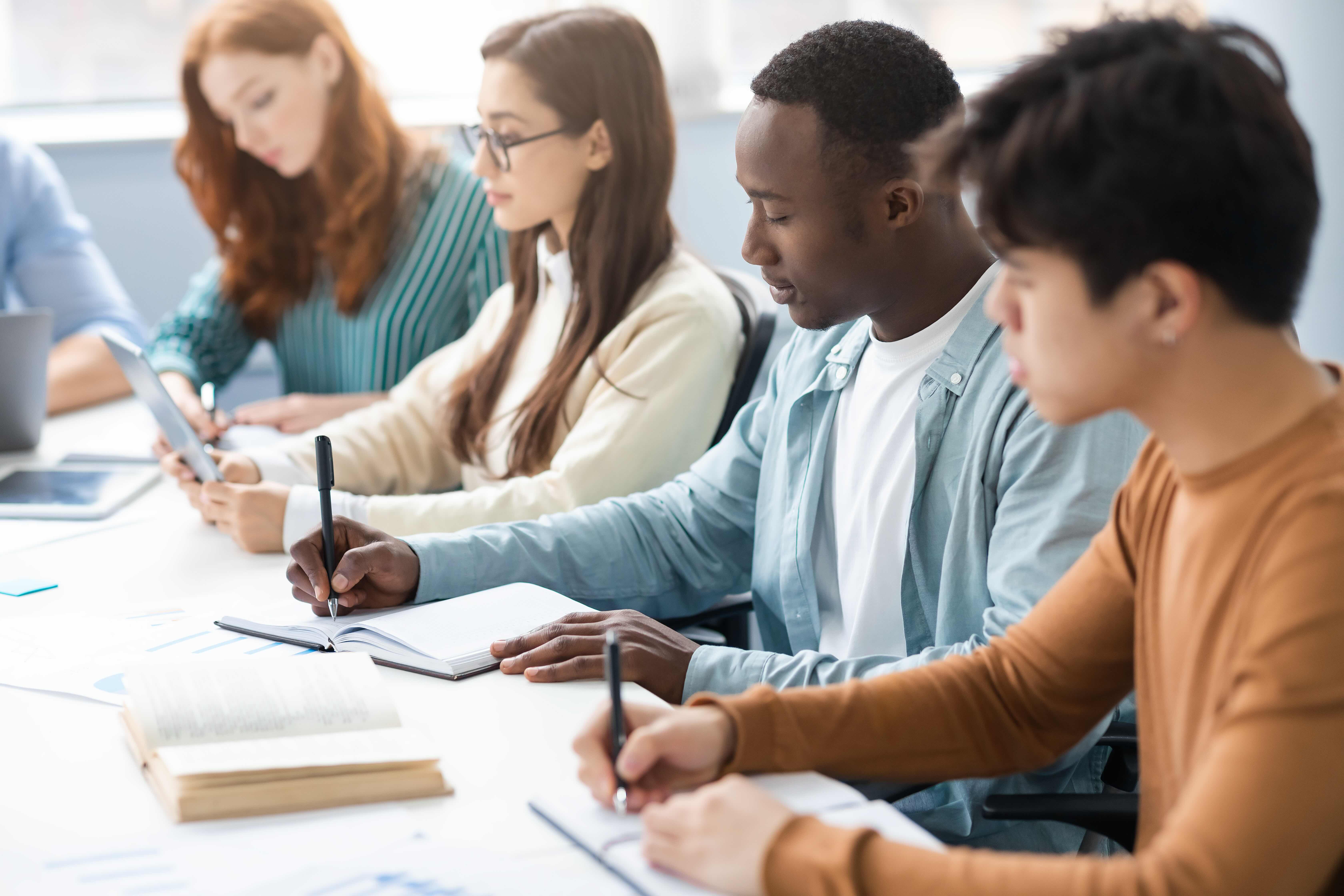 Diverse Students Sitting At Desk In Line Taking Notes