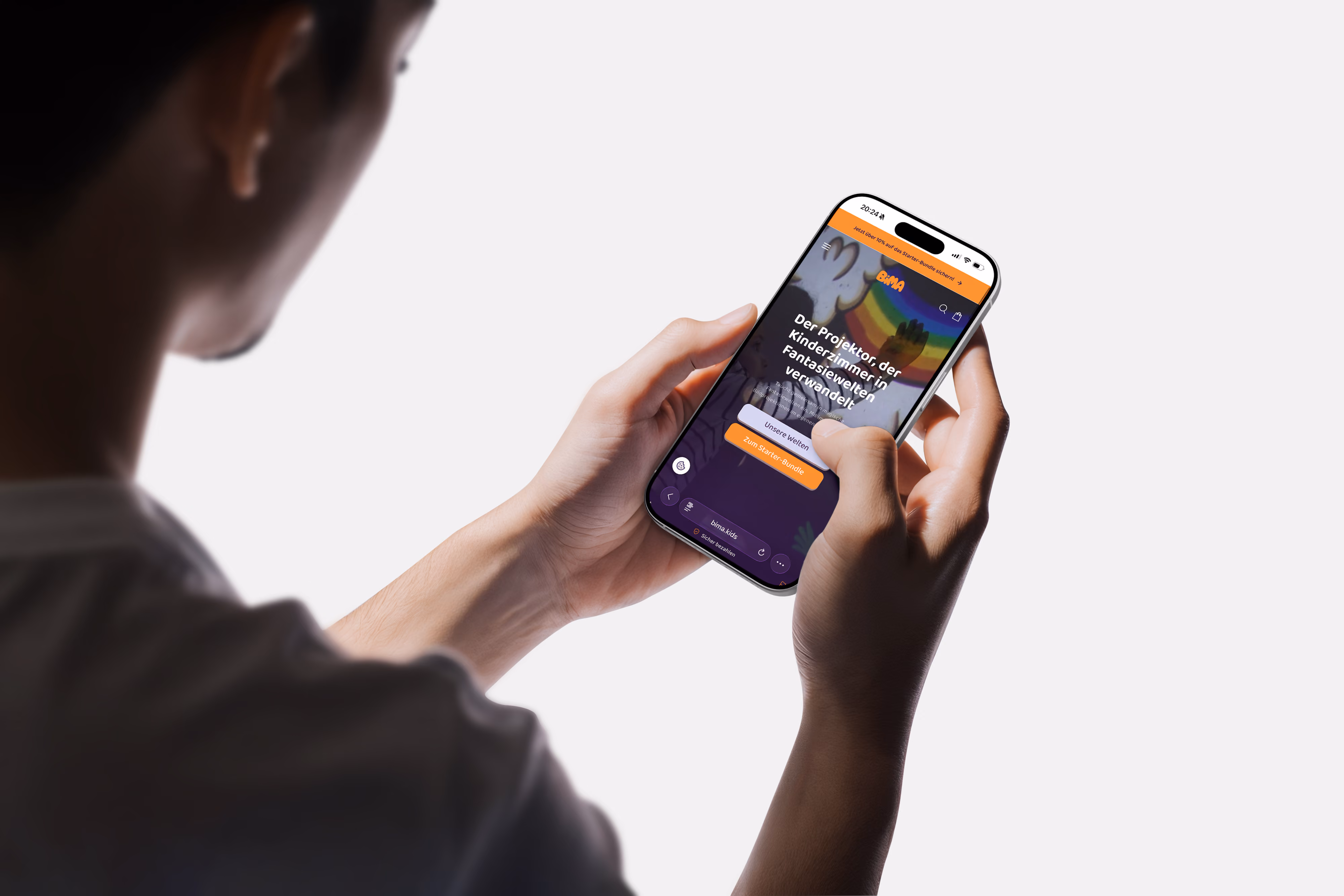 Person hält ein Smartphone mit der Website von BIMA, die einen Projektor zeigt, der Kinderzimmer in Fantasiewelten verwandelt. Der Bildschirm zeigt Buttons für 'Unsere Welten' und 'Zum Starter-Bundle'.