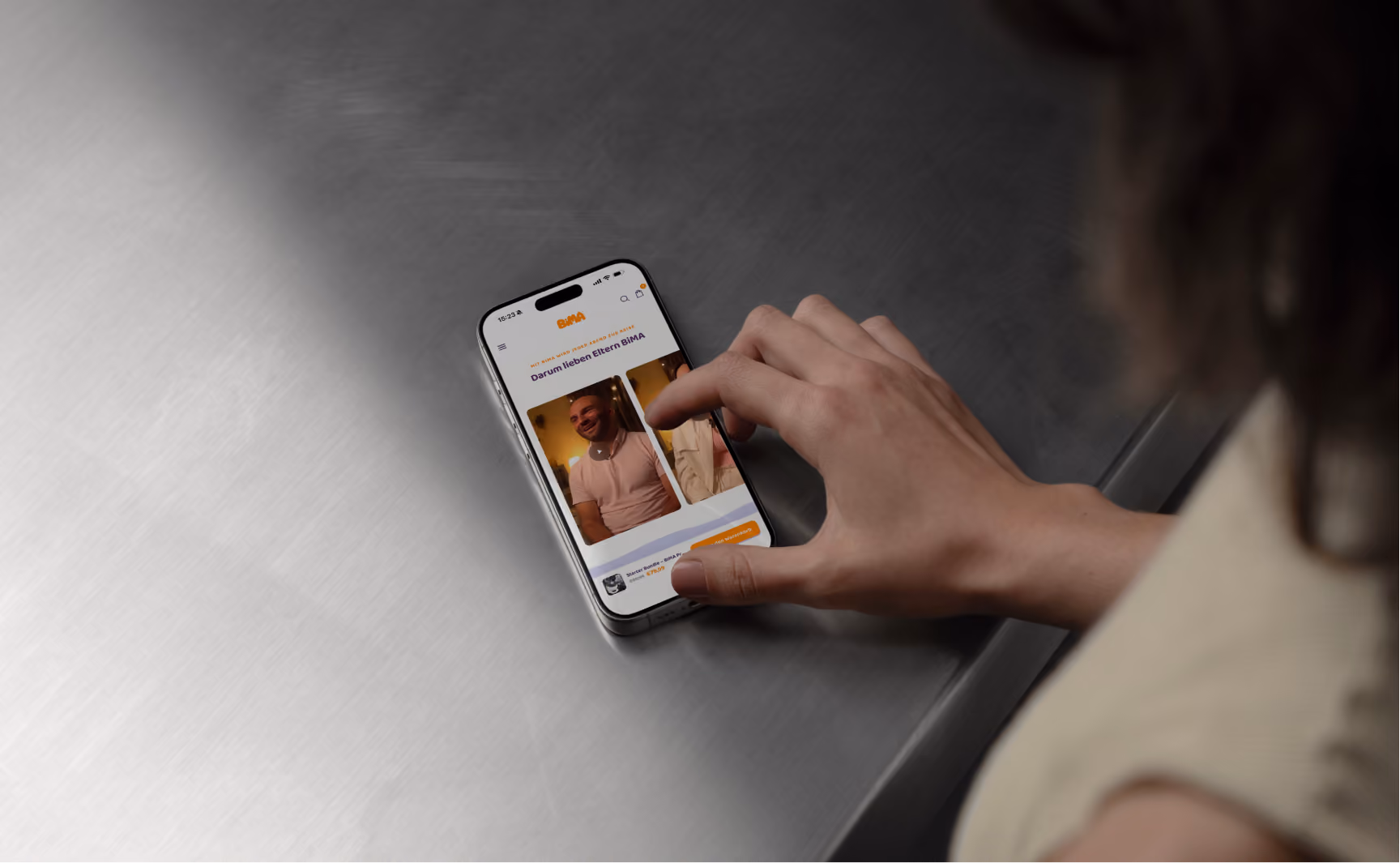 Eine Hand bedient ein Smartphone auf einem Metalltisch. Der Bildschirm zeigt eine Webseite mit dem Titel 'Darum lieben Eltern BiMA' und zwei Videos mit Personen, dazu ein orangefarbener Button unten rechts.