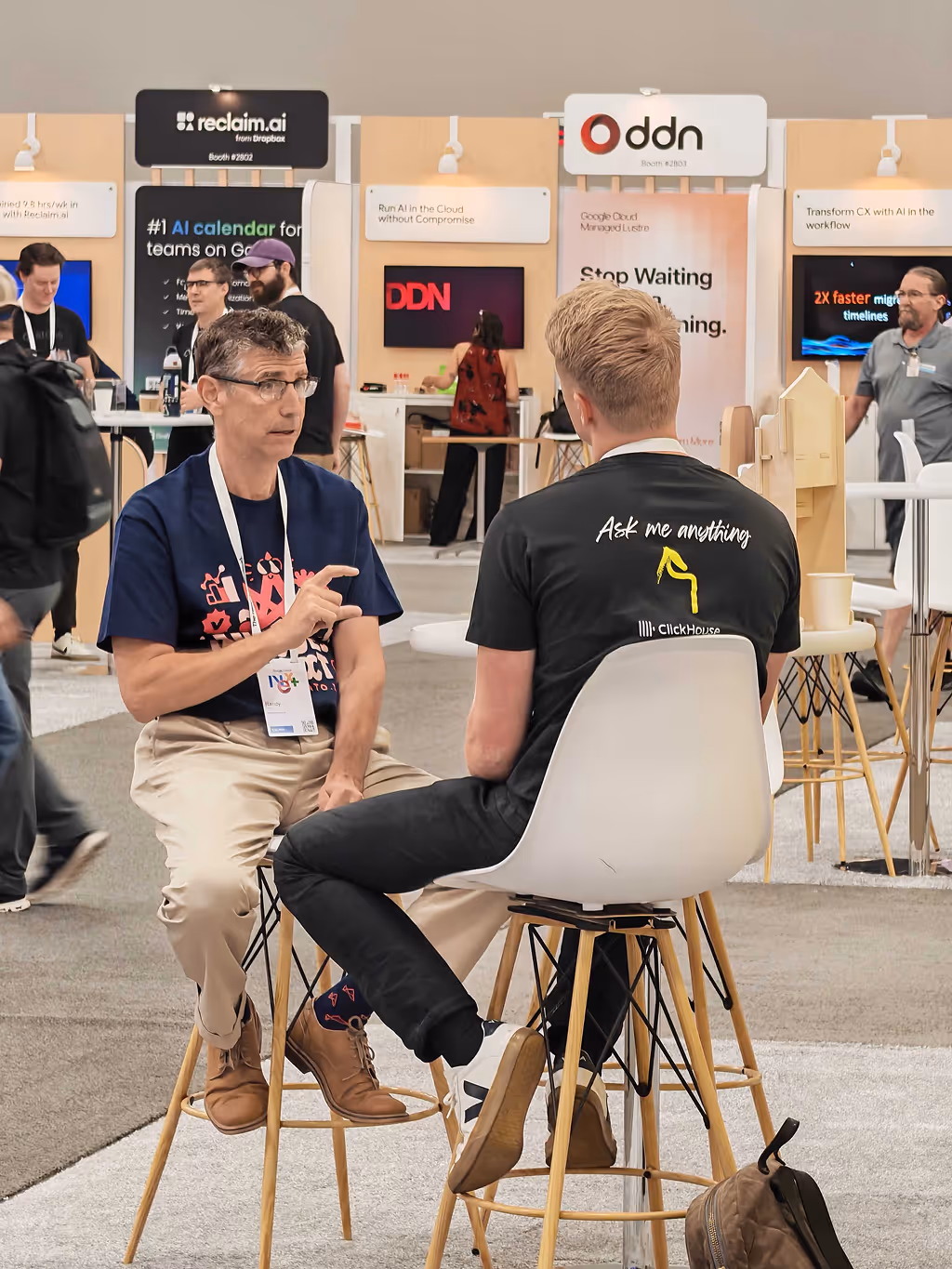 Two men seated on stools having a conversation at a tech conference with exhibition booths in the background.