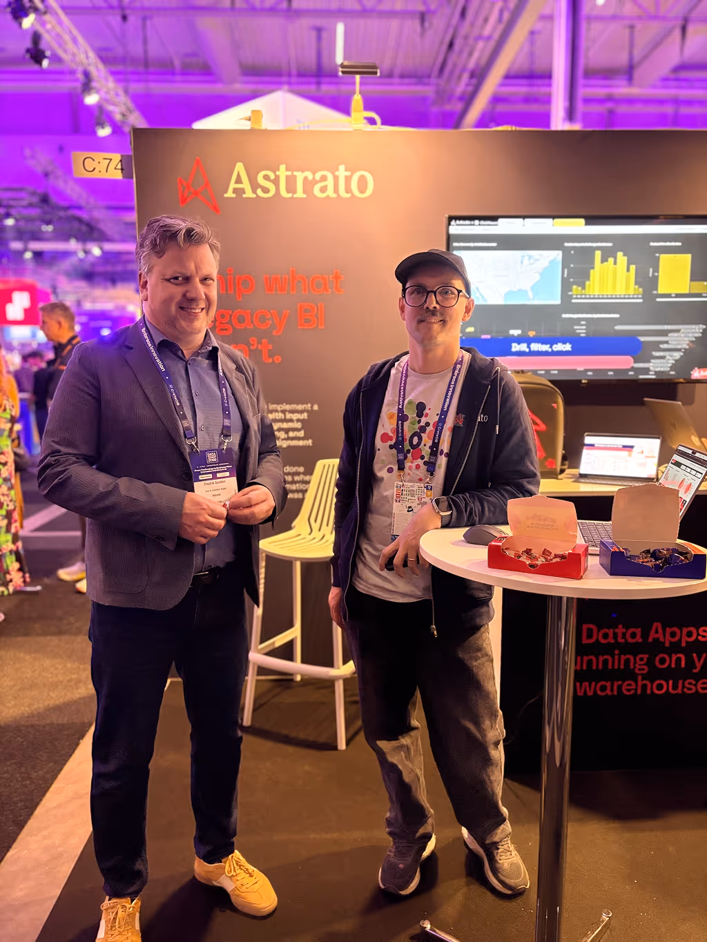 Two men standing in front of an Astrato booth with data charts on screen and candy boxes on a round table.