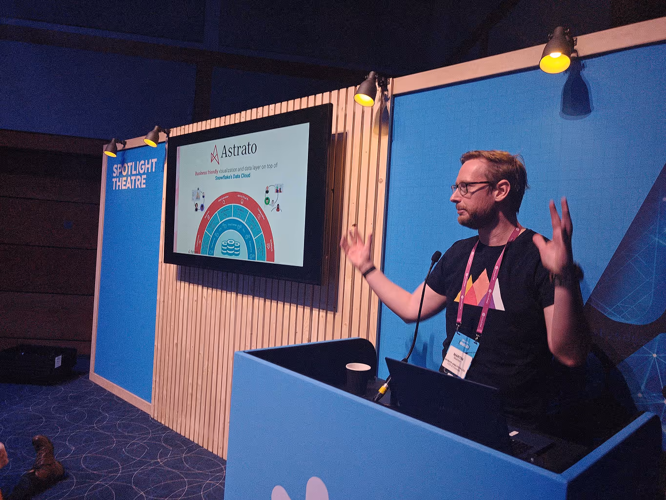Man with glasses giving a presentation at Spotlight Theatre with a display showing Astrato business visualization.