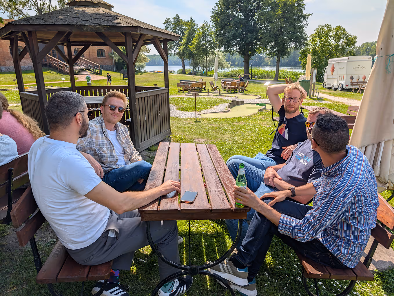 Five men sitting at a wooden picnic table outdoors near a gazebo, drinking and chatting on a sunny day.