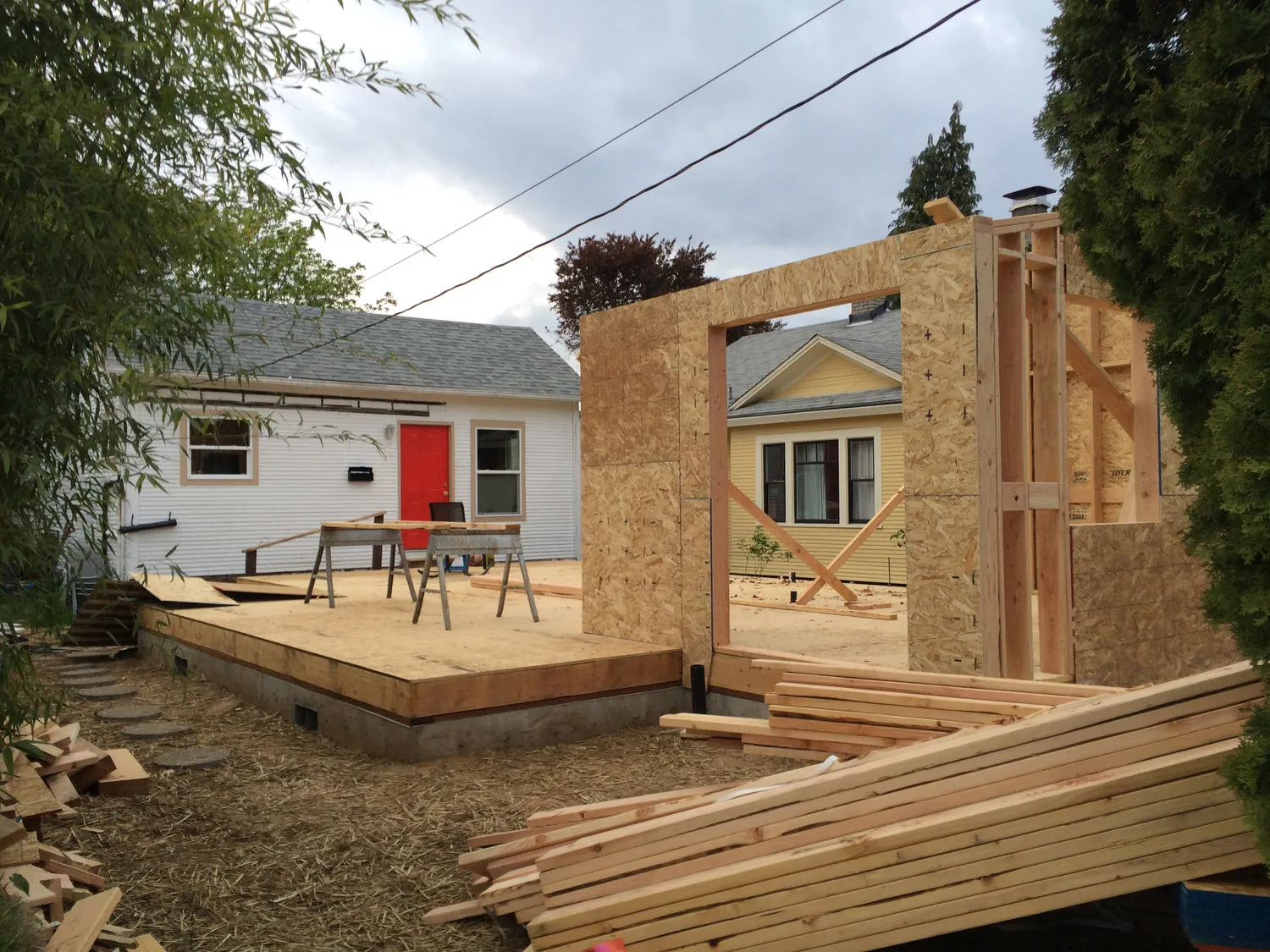 Partially constructed wooden house frame with stacked lumber in the foreground and neighboring houses in the background.