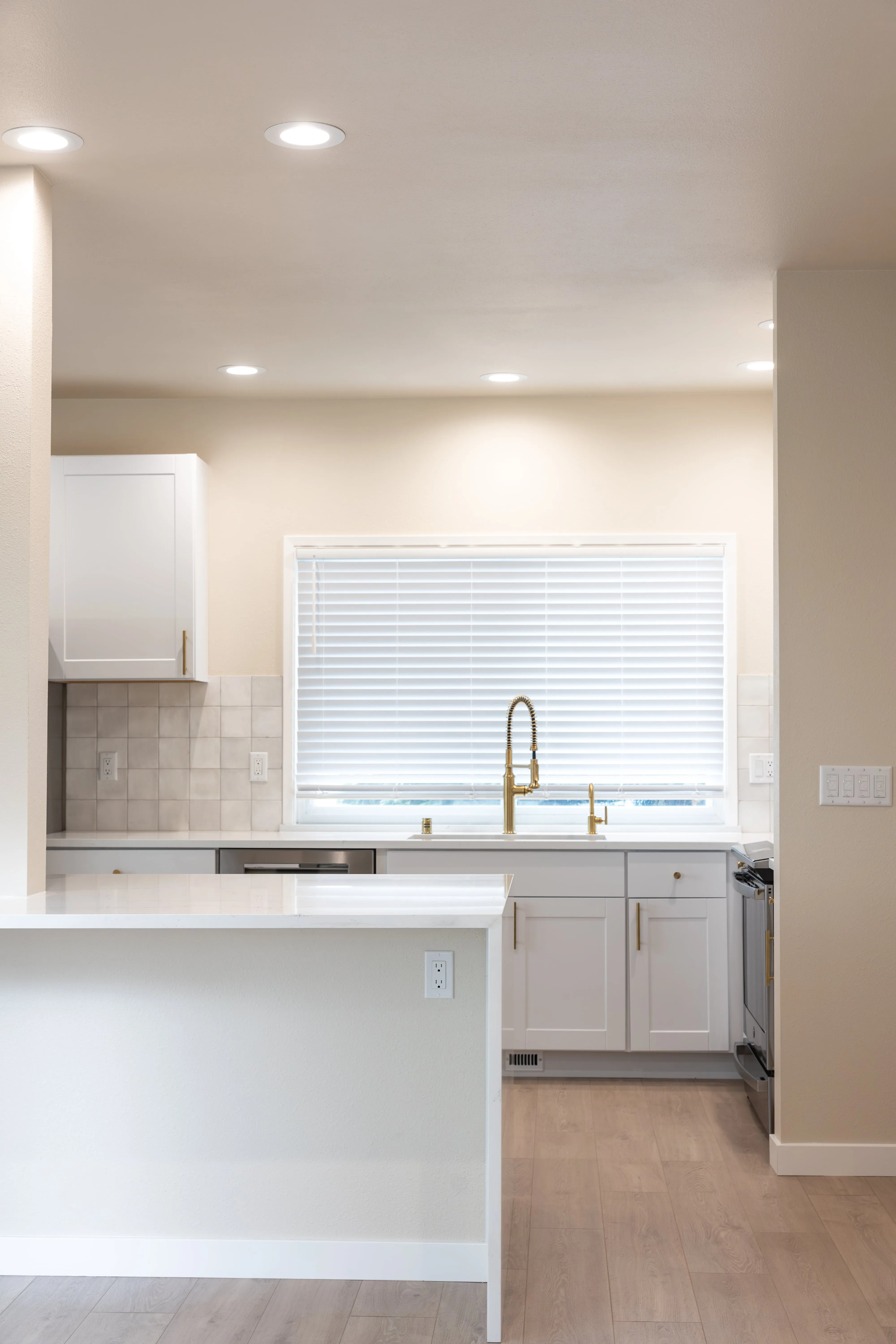 Modern kitchen with white cabinetry, brass faucet, large window with blinds, and light wood flooring.