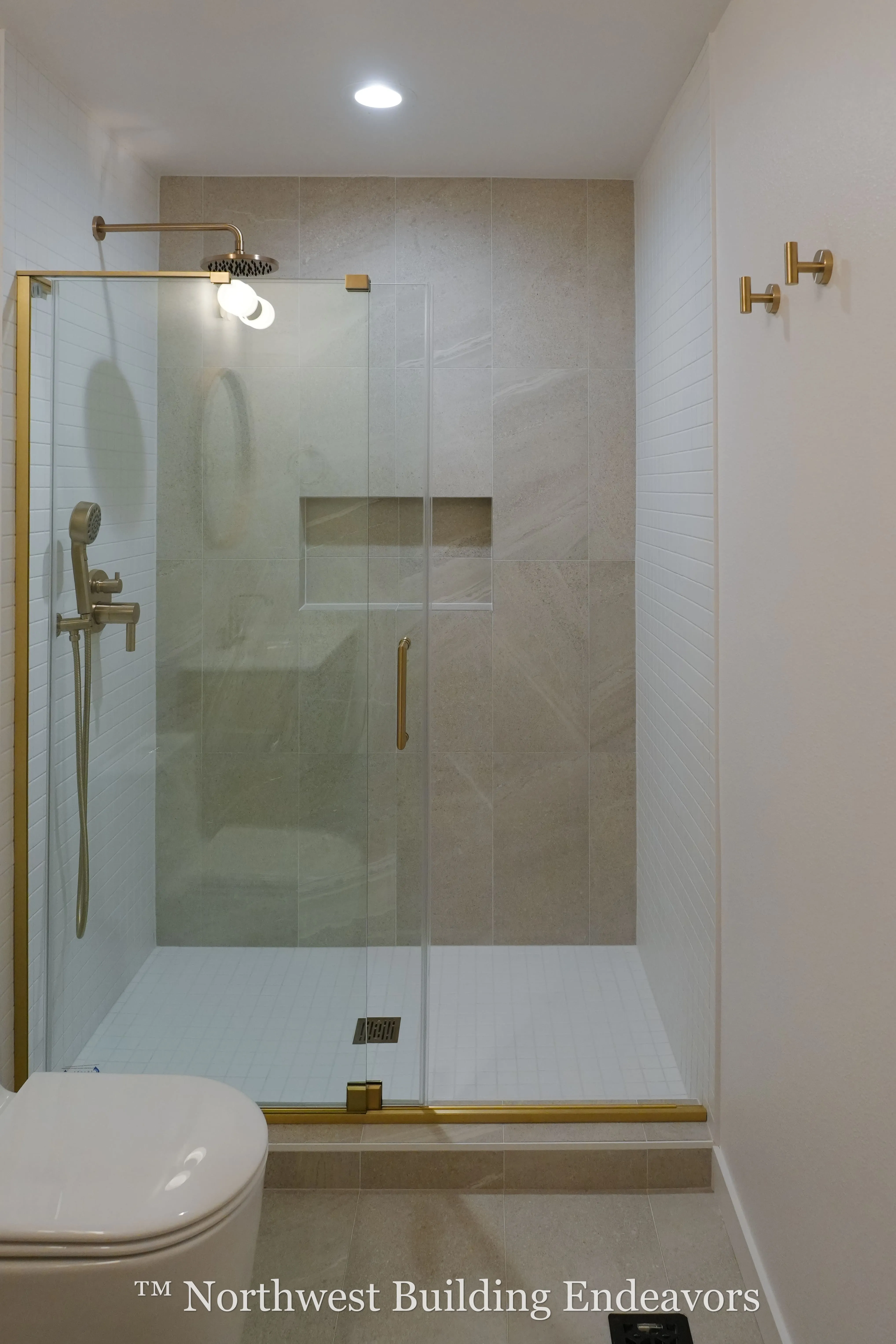 Modern bathroom shower with beige tile and gold fixtures, including rainfall and handheld showerheads, enclosed by glass door.