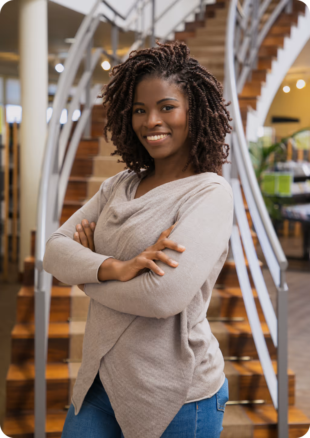 Confident woman with curly hair smiling with arms crossed in front of modern indoor staircase.
