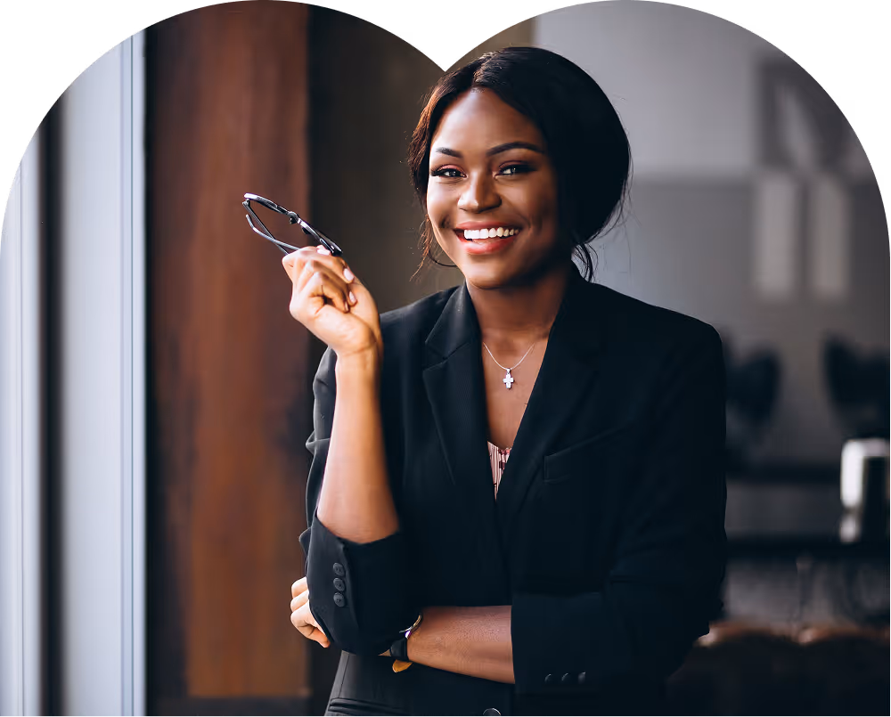 Smiling professional woman in a black blazer holding glasses near a window in an office.