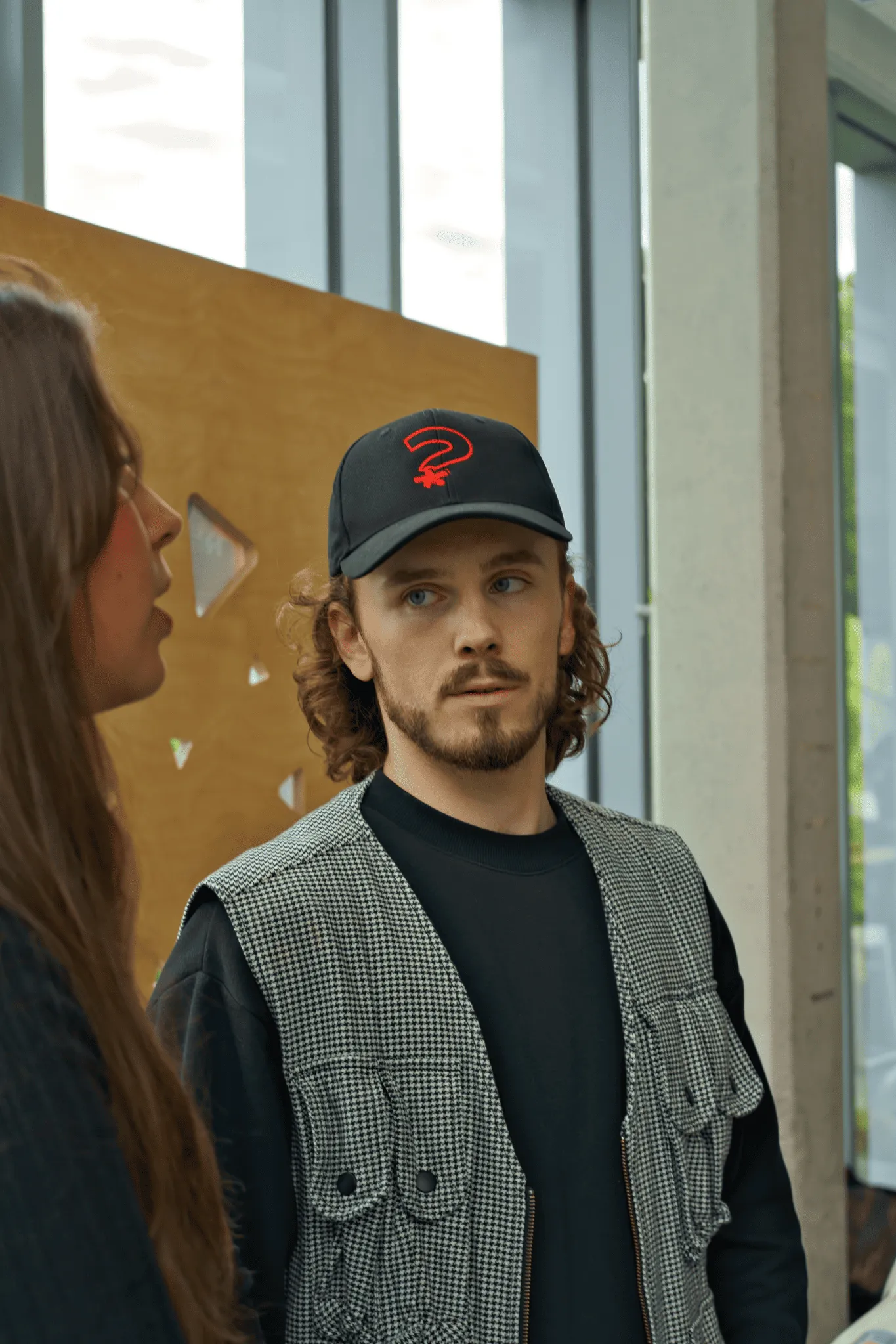 Un homme portant une casquette noire avec un point d'interrogation rouge discute avec une femme dans un intérieur lumineux moderne.