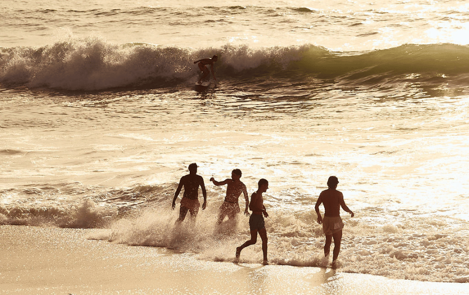 A group of friends is walking into the waves, while a surfer rides a wave in the background.