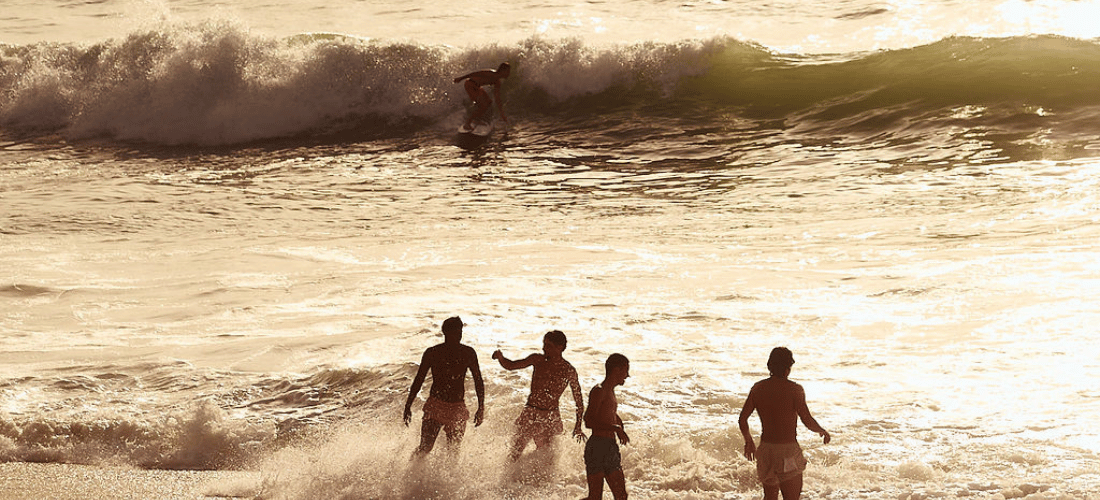 Four people are splashing in the shallow water at the beach. Further out, one person is surfing on a wave under a bright, golden sky.