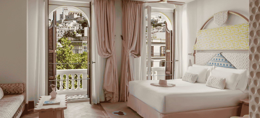 Une chambre spacieuse de style méditerranéen, dont les portes-fenêtres s'ouvrent sur une vue imprenable sur une ville côtière historique.
