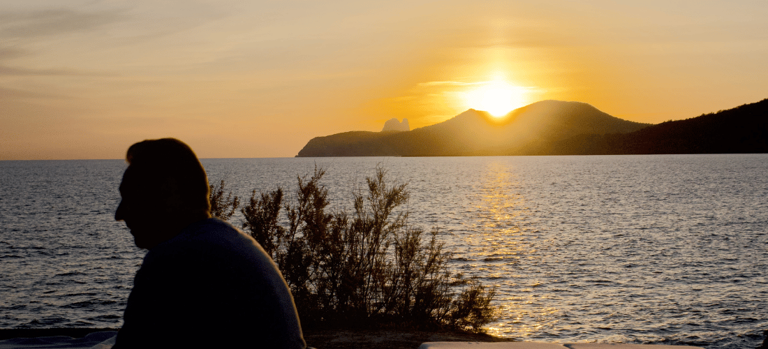 Silhouette d'une personne assise au bord de la mer au coucher du soleil, avec des transats vides à proximité et des collines au loin.