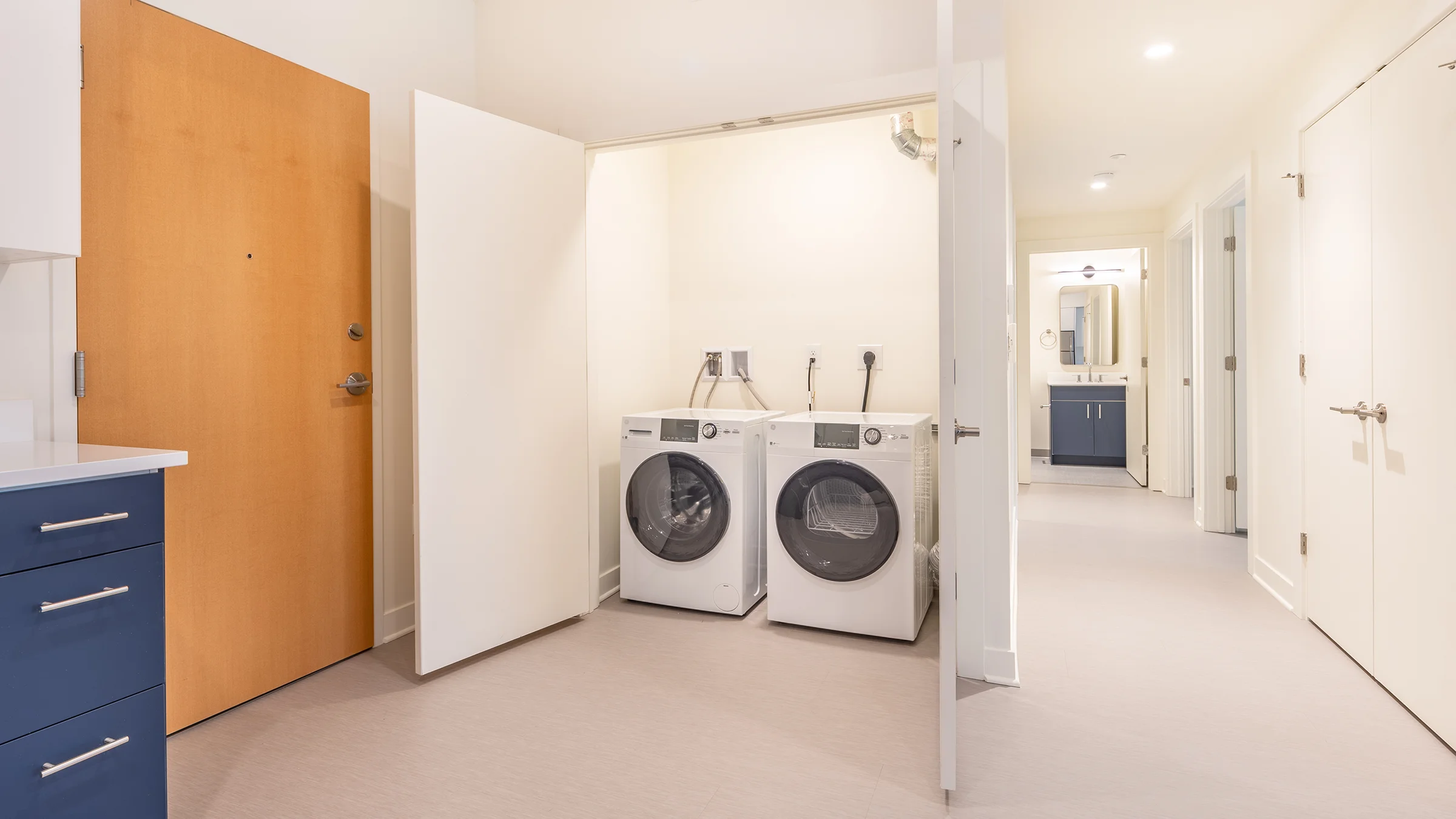 Laundry facilities and hallway to bathroom in ADA compliant apartment. White walls, light floor and blue cabinetry make for an elevated but clean space.
