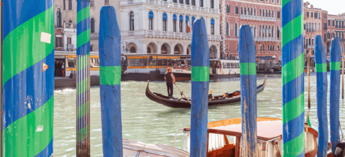 Venice Port, Italy – A man navigating a traditional gondola boat along a scenic waterway in Venice, surrounded by the city's iconic canals and waterfront views.