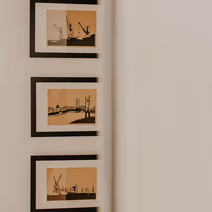 Trois illustrations encadrées représentant des scènes portuaires stylisées, accrochées sur un mur blanc, partiellement vues à travers une porte.