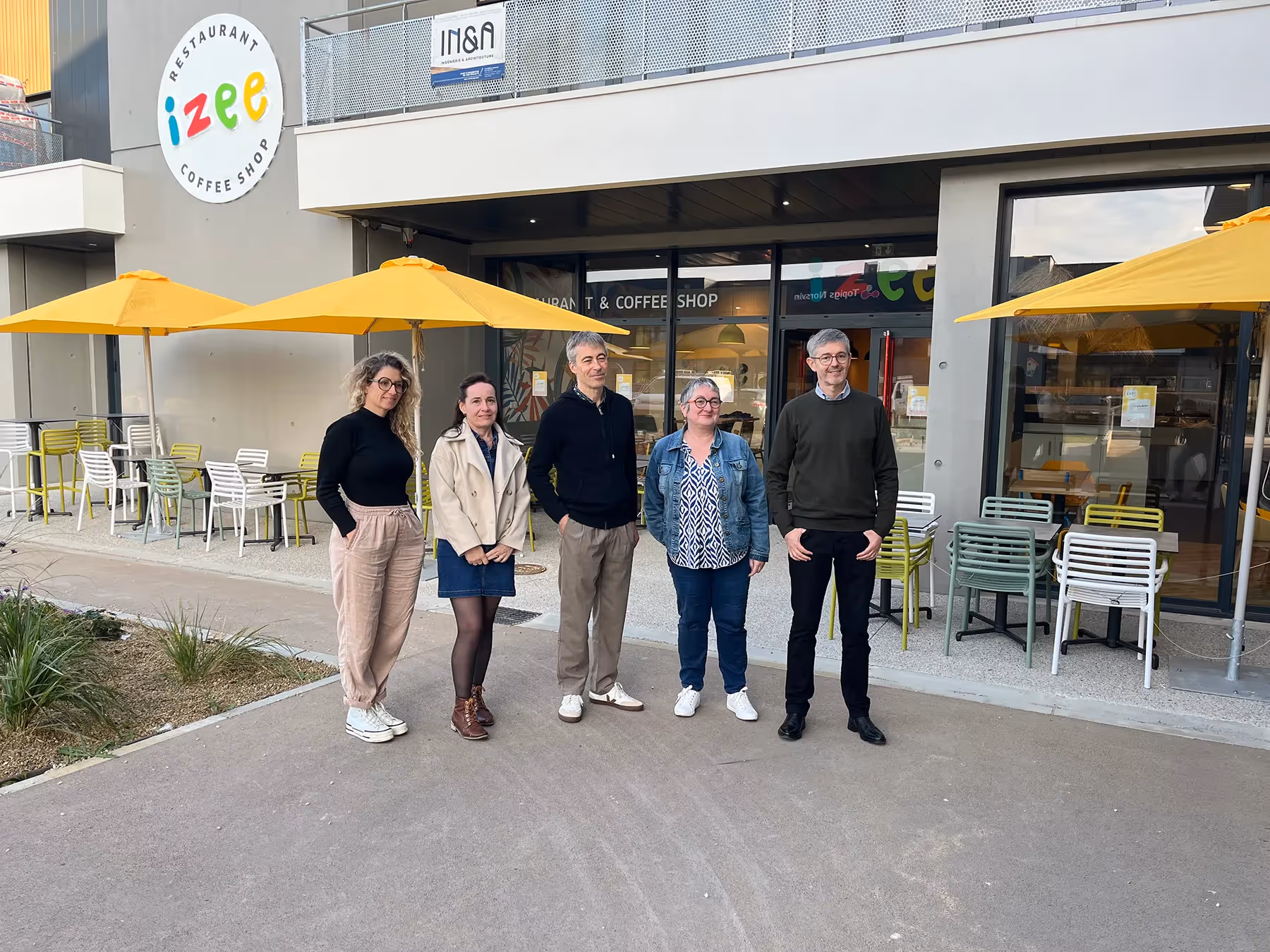 Cinq personnes posent devant le restaurant coffee shop Izee avec terrasses et parasols jaunes.