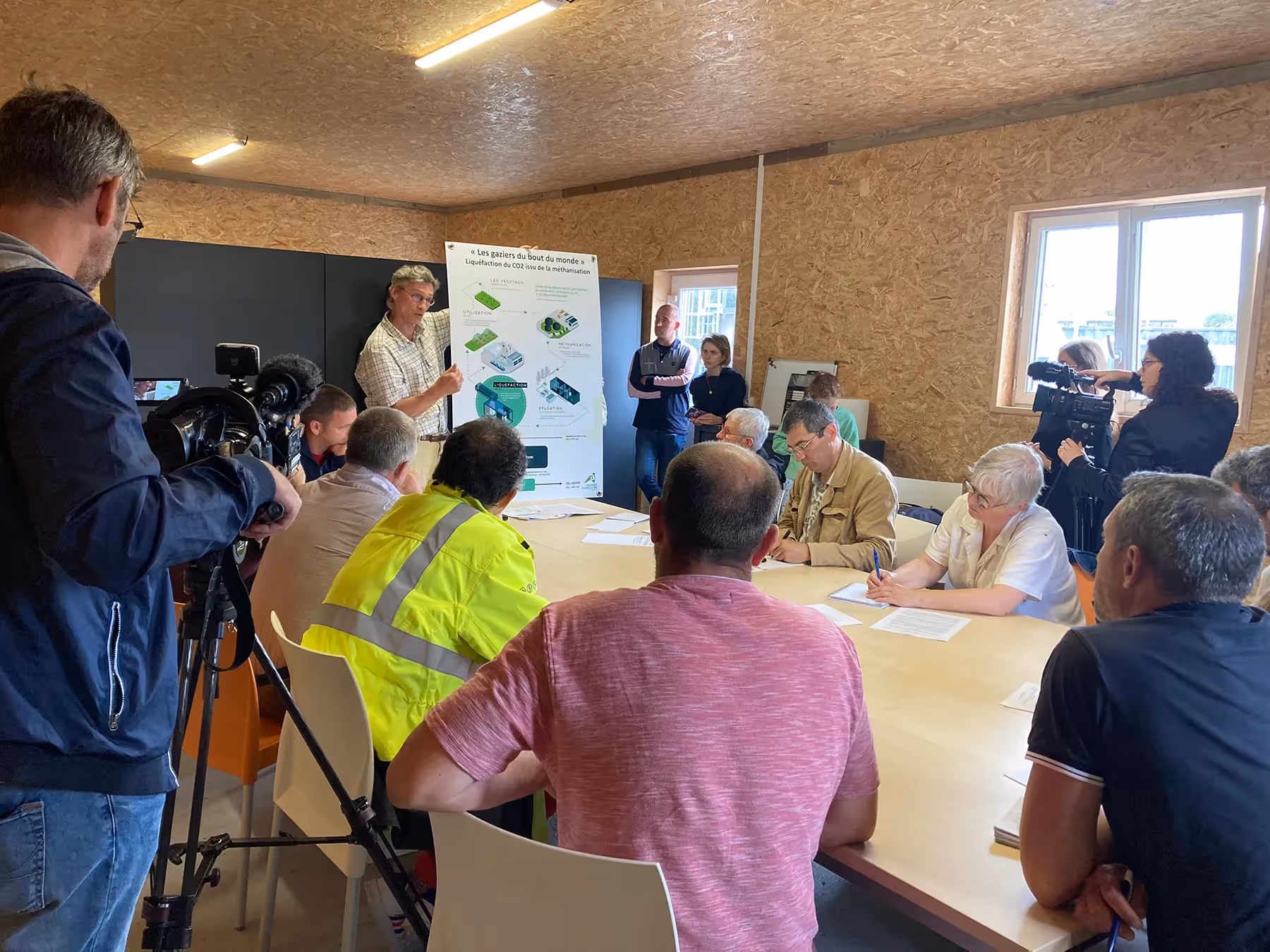 Réunion d'information autour d'une table avec présentation de projets écologiques et présence de caméras.