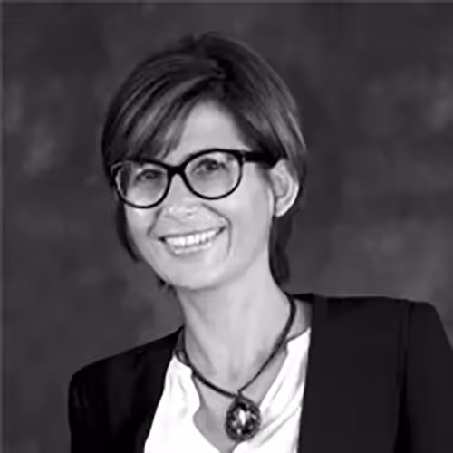 Portrait professionnel en noir et blanc d’une femme souriante portant des lunettes et un collier, sur fond studio.