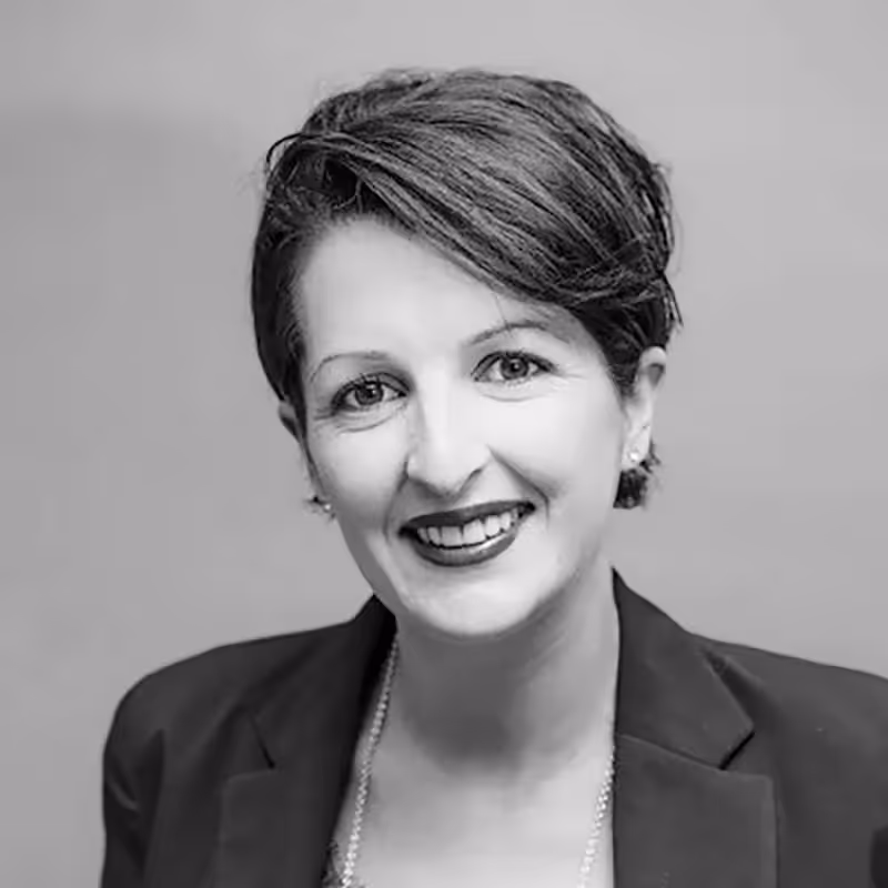 Portrait professionnel d’une femme souriante en noir et blanc, cheveux courts et tenue élégante, sur fond uni.