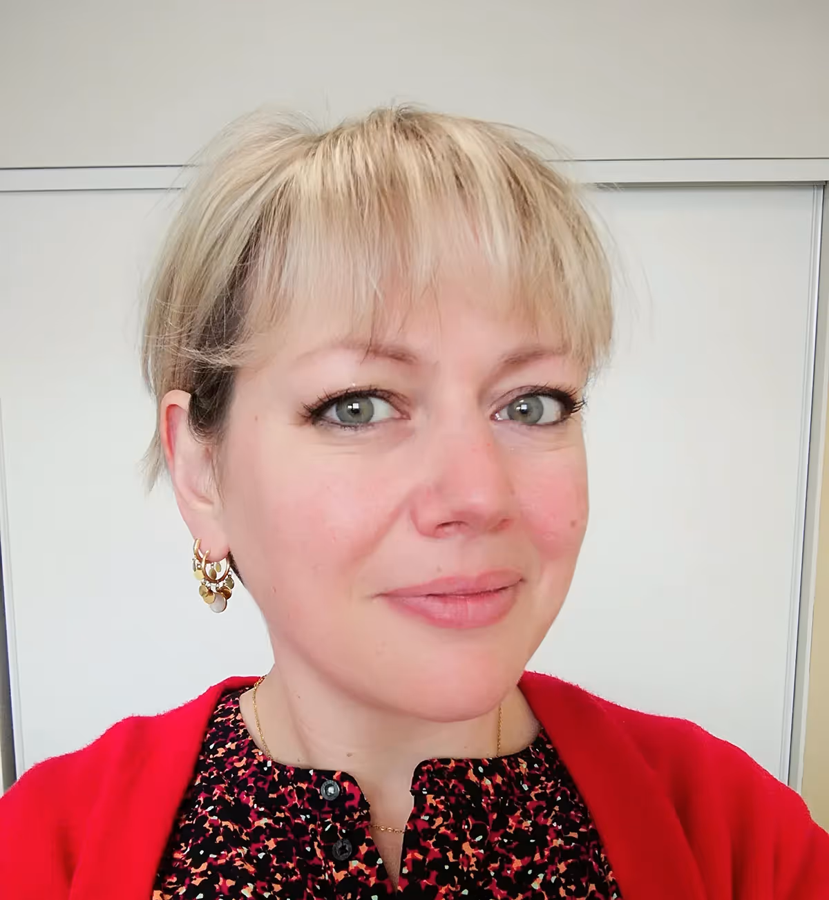 Portrait d’une femme souriante aux cheveux blonds courts, vêtue d’un haut à motifs colorés et d’un gilet rouge, posant en intérieur sur fond clair.