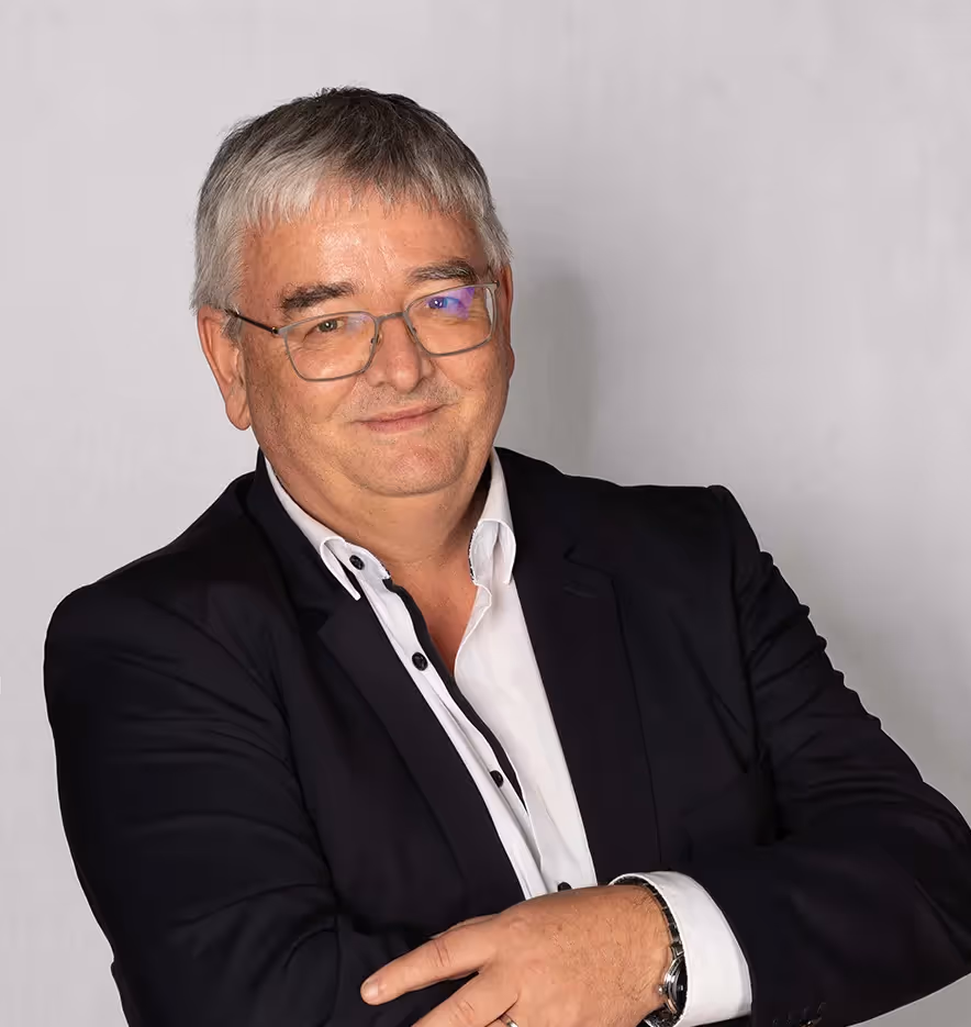 Homme aux cheveux gris en costume foncé et chemise blanche, posant avec assurance sur fond neutre.