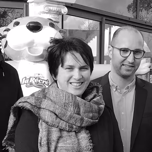 Deux personnes souriantes posent devant une mascotte de parc d’attractions portant l'inscription "La Récré des 3 Curés".