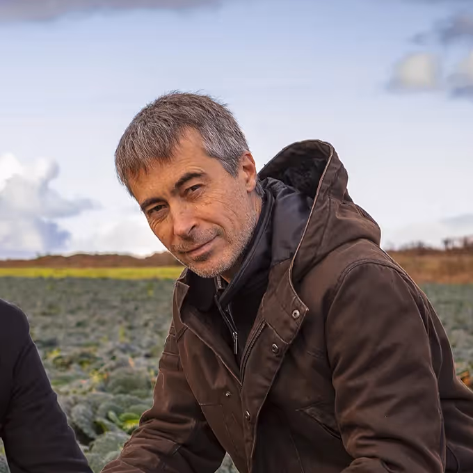 Homme en manteau brun dans un champ de légumes, regardant la caméra avec un léger sourire sous un ciel nuageux.