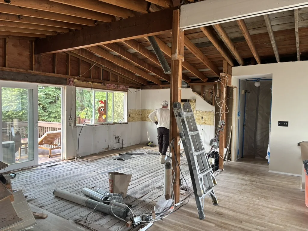 Room with exposed ceiling joists, partially removed drywall, ladder, ductwork, and sliding glass doors leading to a deck