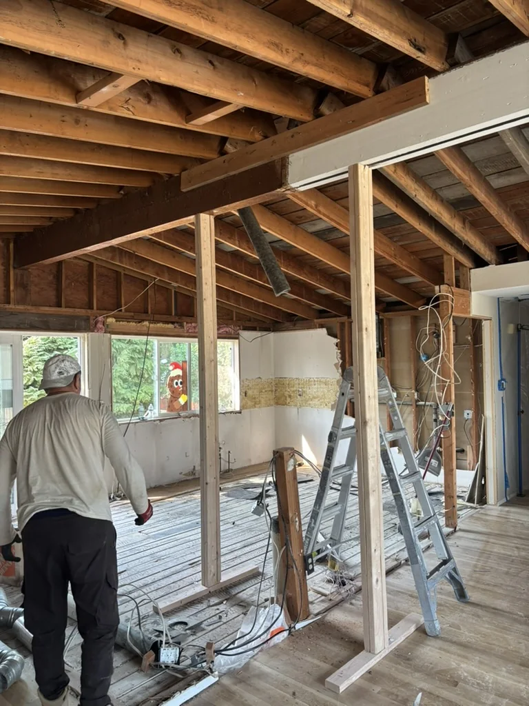 Interior room with exposed ceiling joists, stripped walls, wooden floorboards, ladders, hanging wires, and a person in a cap walking through the space.