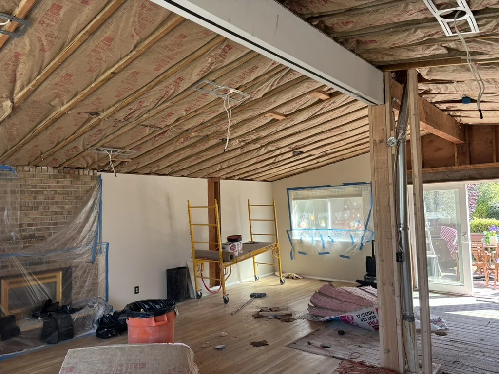 Room with exposed ceiling framing, plastic sheeting, a yellow scaffold, and a bucket on the floor