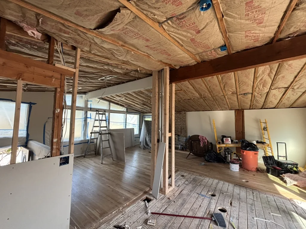 Room under construction showing exposed wood ceiling joists, insulation, framed walls, ladders, drywall sheets, and scattered tools on plank flooring.