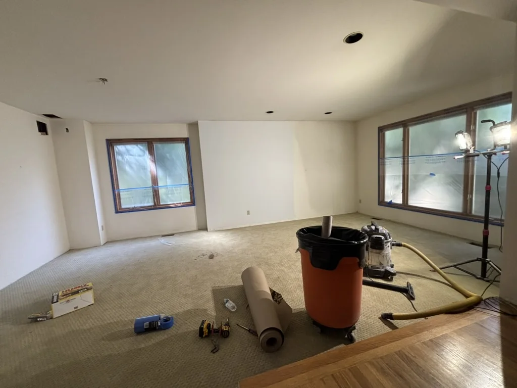 Empty room with beige carpet, white walls, windows covered in protective film, and tools on the floor