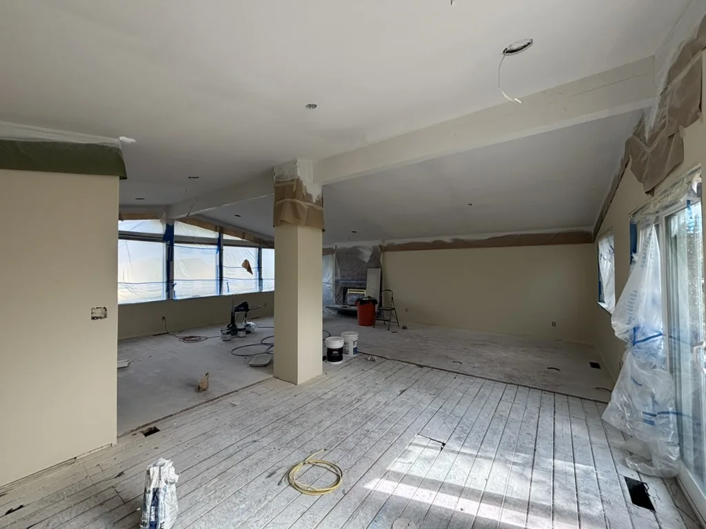 Large room with exposed ceiling, plastic-covered windows, construction tools, paint buckets, and unfinished wooden flooring