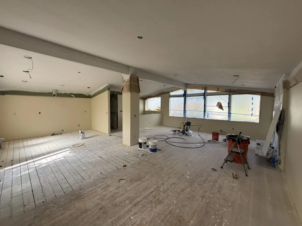 An unfinished room with beige walls, exposed wood subfloor, plastic covered windows, paint buckets, and a step ladder
