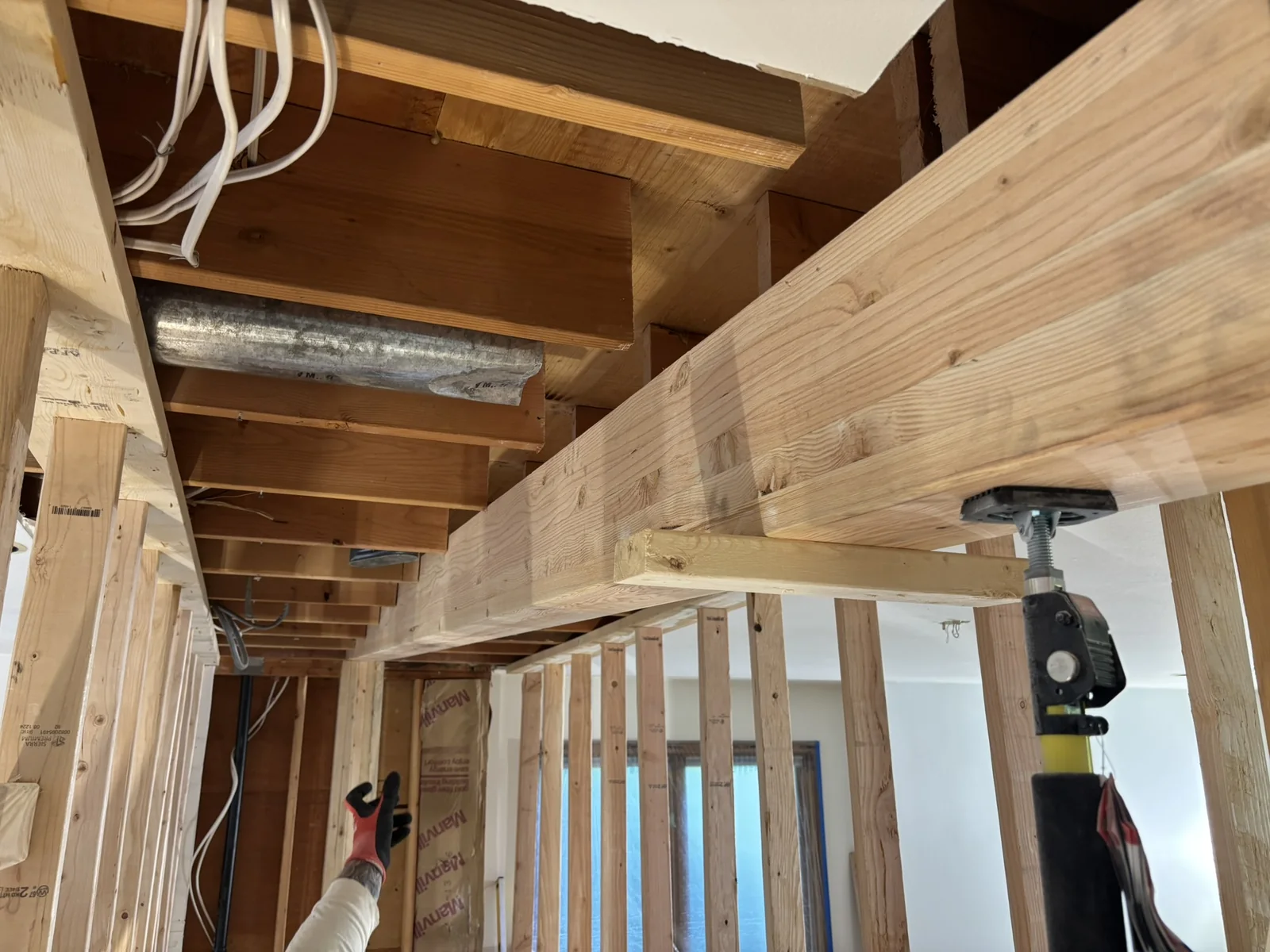 A large wooden beam spans the ceiling above vertical wall studs, metal ductwork, and an adjustable steel support post.