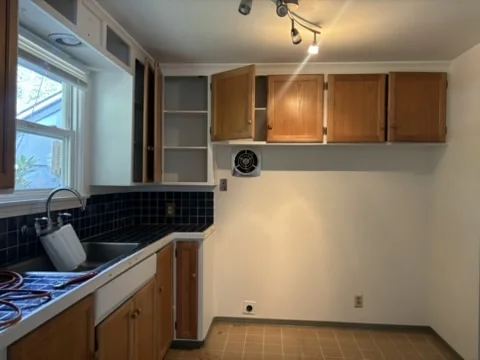 Wooden kitchen cabinets, sink with faucet, blue tile backsplash, window, and tiled floor under track lighting.