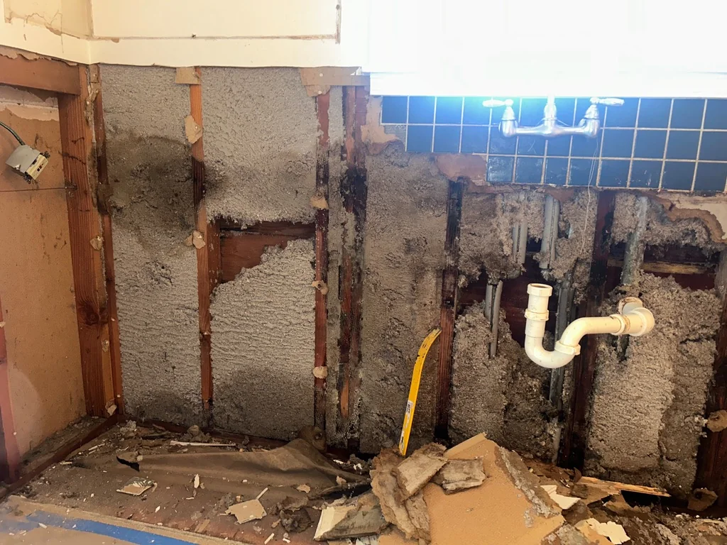 A partially demolished interior wall reveals wooden studs, insulation, a white PVC pipe, and blue tiled backsplash with a faucet.